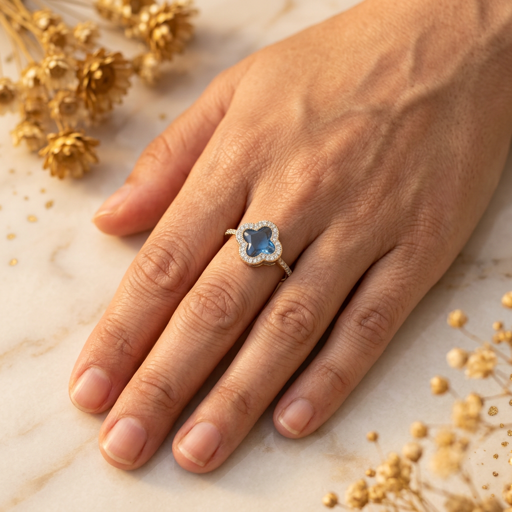 Adult hand wearing a blue quatrefoil clear-stone halo ring while resting naturally on cream marble.