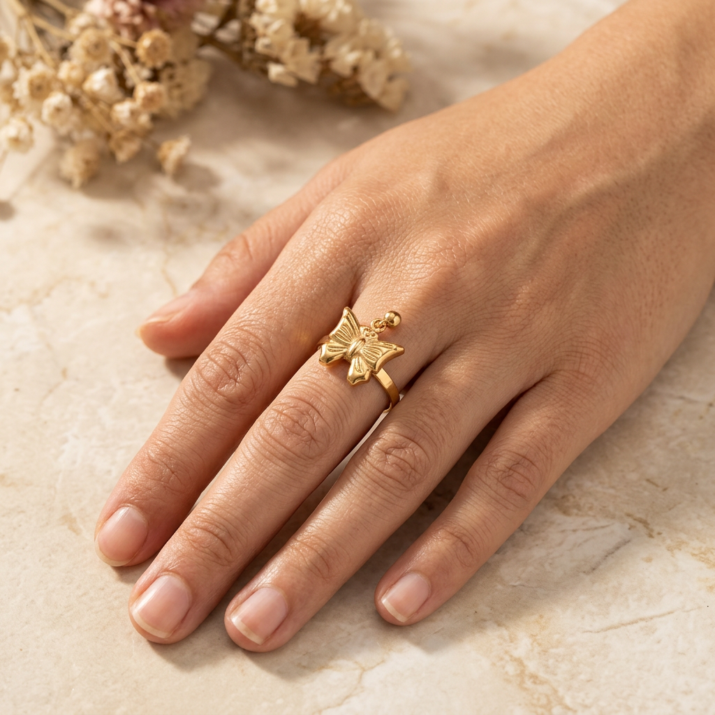 Adult hand wearing a gold-tone butterfly ring with bead detail, resting naturally on cream marble.