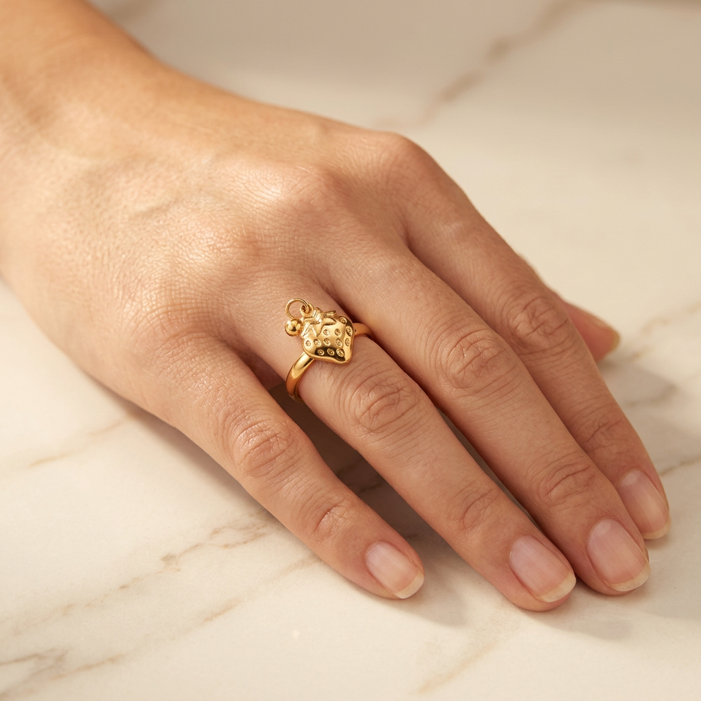 Adult model wearing gold-tone strawberry ring with hand resting on cream marble.