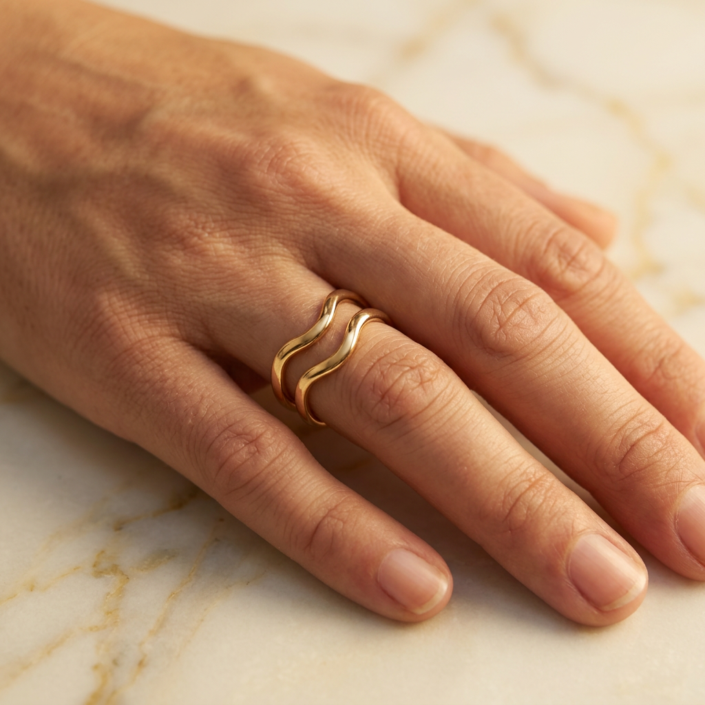 Adult model wearing gold-tone double-wave ring with hand resting on cream marble.