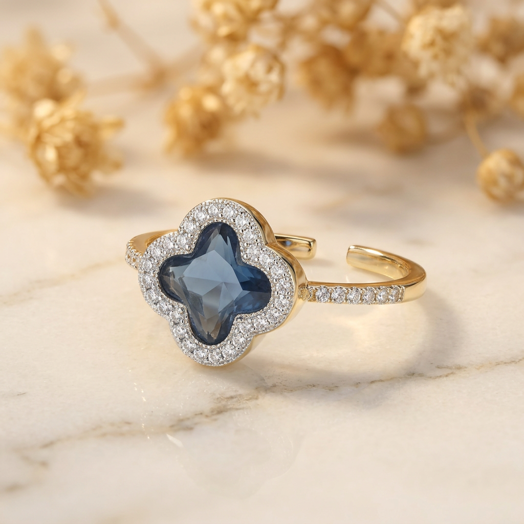 Blue quatrefoil halo ring with clear-stone shoulders on cream marble with blurred golden flowers.