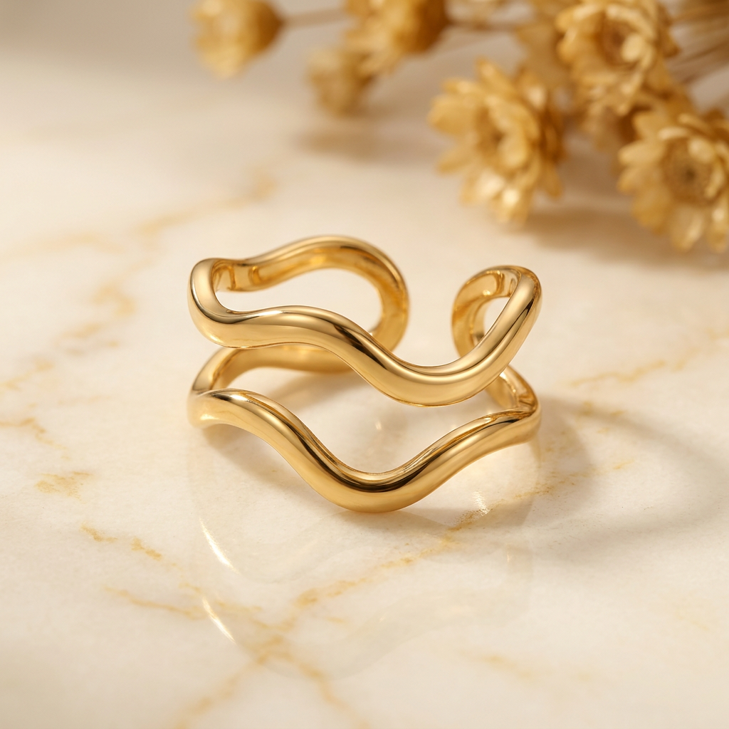 Gold-tone double-wave ring on cream marble with blurred golden dried flowers.
